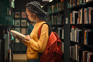 Female college student reads a book among bookcases full of books.
